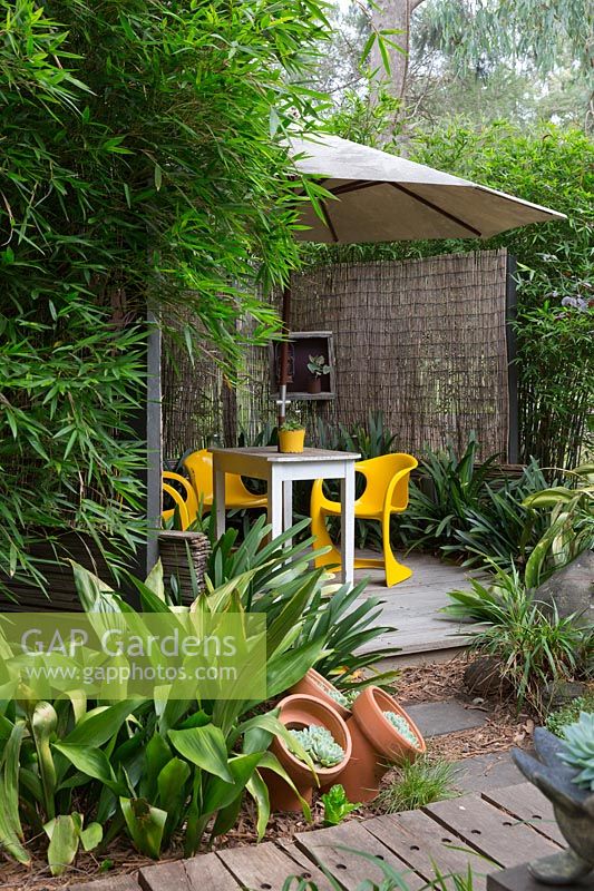 Timber deck covered by a large white umbrella with bright yellow plastic retro chairs and a timber table, screened with bamboo matchstick blinds and framed by a clump of bamboo and Aspidistra elatior, cast iron plant.