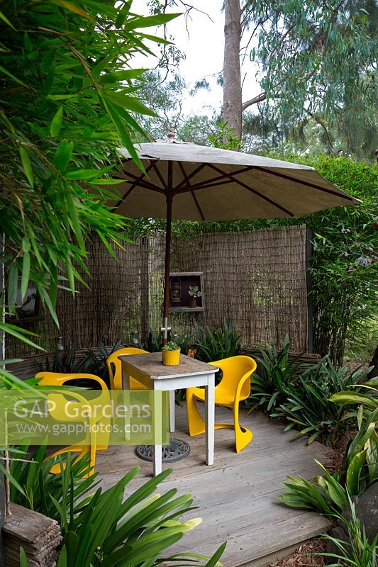 Timber deck covered by a large white umbrella with bright yellow plastic retro chairs and a timber table, screened with bamboo matchstick blinds and framed by a clump of bamboo andAspidistra elatior, cast iron plant.