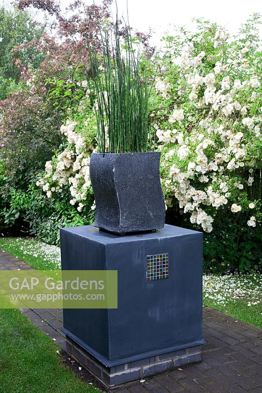 Grey planter filled with Equisetum japonicumon - Horsetail - on a monolith with mosaic inset on brick pathway, backed with white rambling roses.