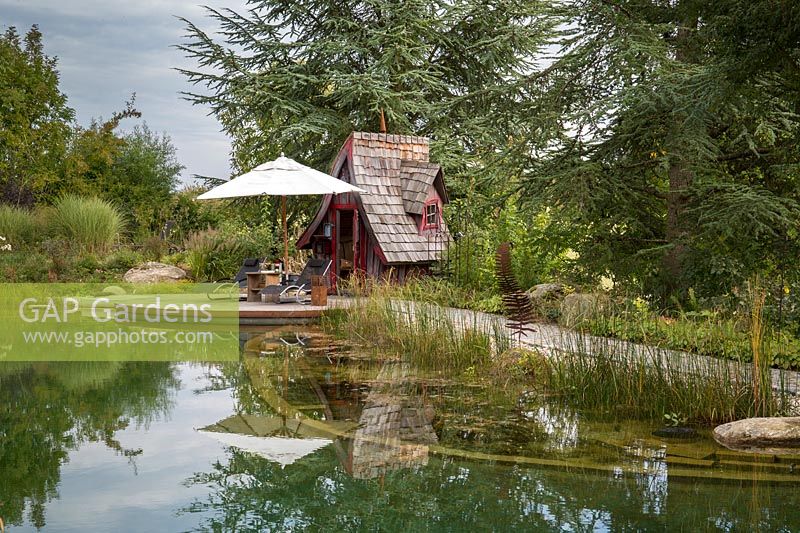 View towards tiny witches' cottage and wooden deck with umbrella on the edge of the natural swimming pond with a zone of clarifying water plants. Miscanthus sinensis 'Morninglight'