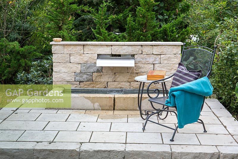 On a flagstone paved area, bistro furniture next to modern water feature with stone wall and water basin