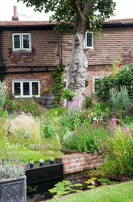 A small low maintenance modern cottage garden planted with self seeding annuals and  perennials. A two tier pond water feature of brick and lead. Lead container filled with Lavender, Stipa tenuissima, Lychnis coronaria, Verbena hastata, Verbena bonariensis.