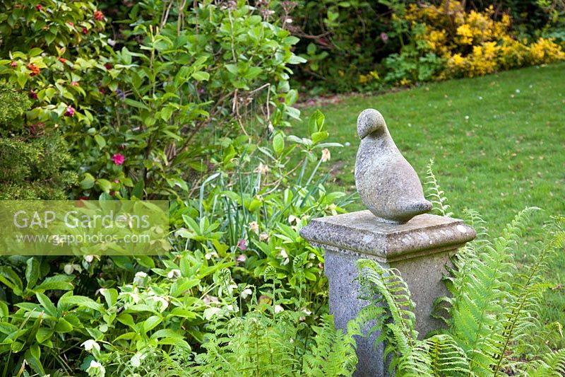 Decorative bird stone statue amongst ferns and helleborus in spring. Garden: Quarry Cottages, Sussex