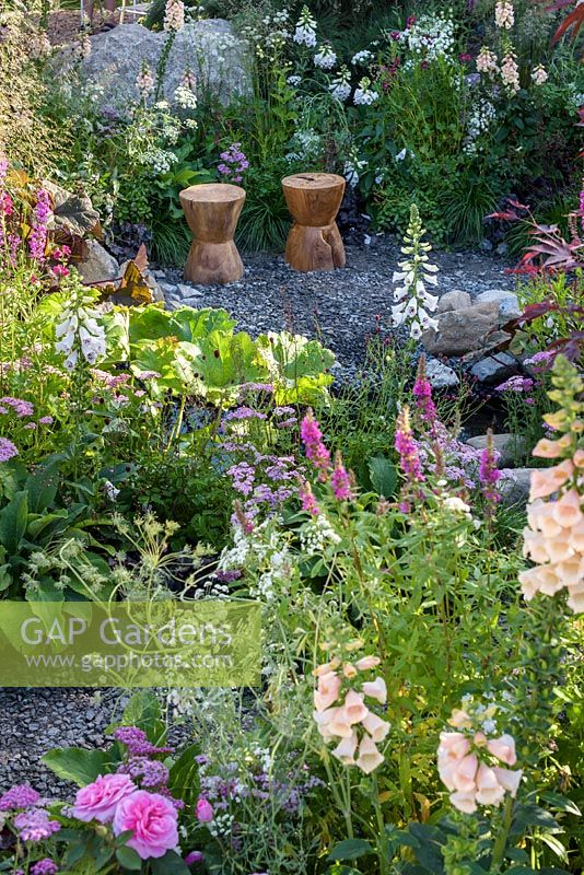 Carved timber seats in gravel area surrounded by deep reds, cream and pink colour themed planting - Great Gardens of the USA, The Oregon Garden, RHS Hampton Court Palace Flower Show 2017. Designer: Sadie May Stowell