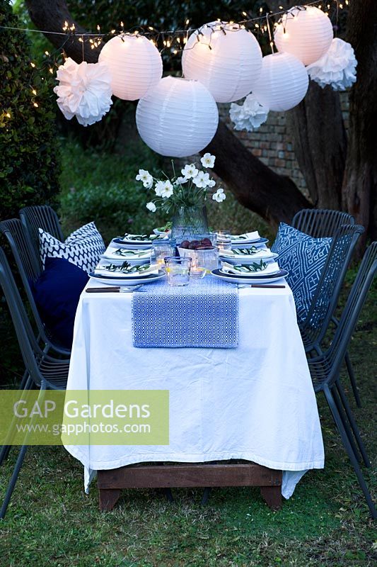 Outdoor dining table dressed in shades of blue and with a vase of white Japanese anemones.  Paper ball lanterns, fairy lights and paper pompoms hang over the table to decorate. Pockets are made in the folded napkins to put name card and each setting is finished with an olive branch. Tealights are put on the table to light it up as dusk falls