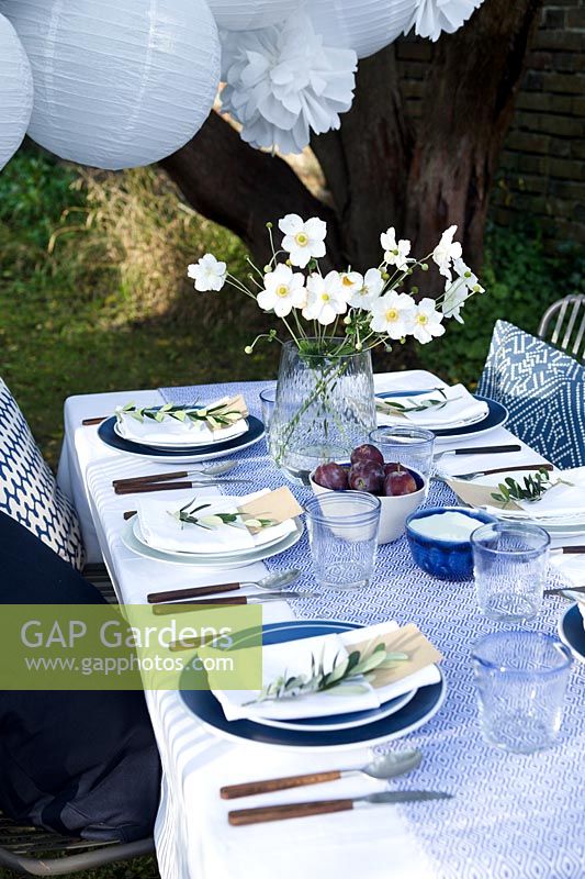 Outdoor dining table dressed in shades of blue and a vase of white Japanese anemones.  Paper ball lanterns and paper pompoms hang over the table to decorate. Pockets are made in the folded napkins to put name card and each setting is finished with an olive branch