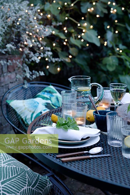 Dusk falls on the outdoor dining area under a fig tree. Fairy lights are in the trees creating a cosy setting and light up the area. Tealights have been lit on the table which is laid with greens and fresh whites and napkins dressed with a geranium leaf