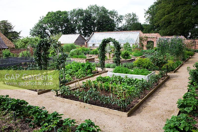 Overview of walled kitchen garden with lean-to greenhouse and Armillary Sphere by David Harber. Raised beds made of drystone walls surrounded by Lavandula 'Imperial Gem'. Steel arches with Apples, Pears and Sweet Peas. Step-over apples and net cages for Brassicas. 
