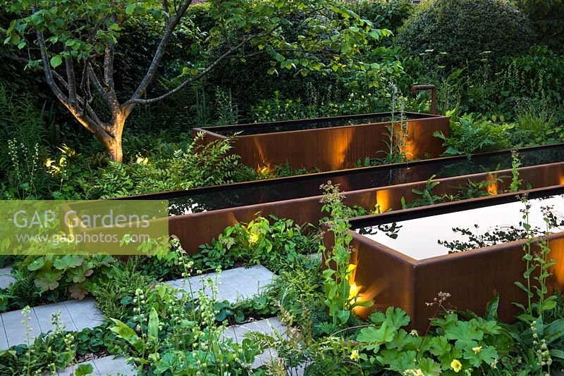 View of three rust steel tanks with water surrounded by soft textural planting and sandstone paving - 'The Zoe Ball Listening Garden' - RHS Chelsea Flower Show 2017 - Designer: James Alexander-Sinclair