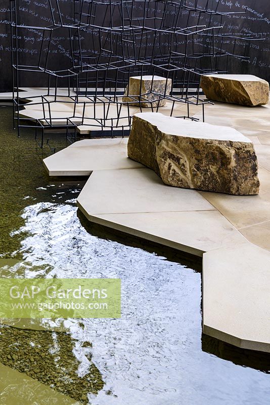 Thin black metal rods sculpture of 'Disappearing Walls' and angular sandstone blocs on fragmented large scale paving overhanging reflective pool - Breaking Ground - RHS Chelsea Flower Show 2017 - Designers: Andrew Wilson and Gavin McWilliam - Sponsor: Darwin Property Investment Management Ltd