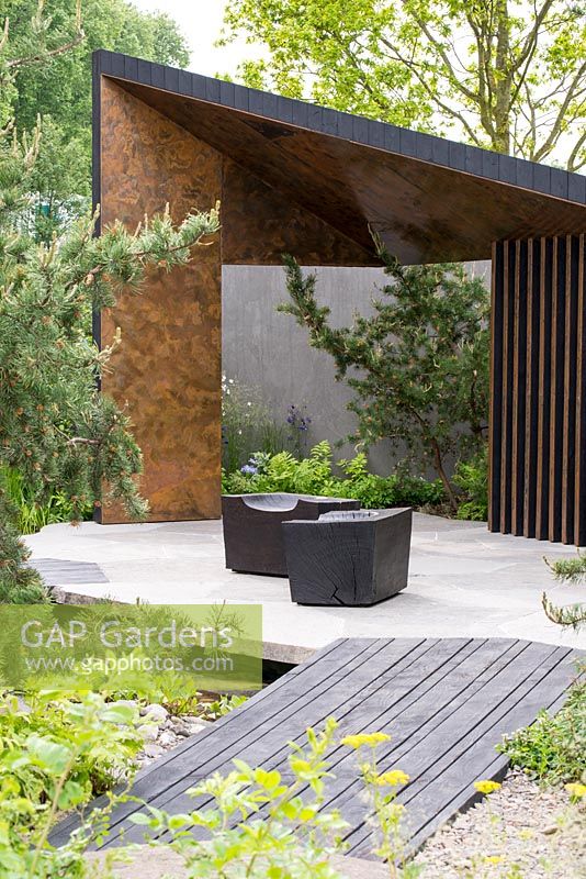 Bridge formed from angular granite blocks leading to a burnt Larch Loggia with cubed seating - The Royal Bank of Canada Garden - RHS Chelsea Flower Show 2017 - Designer: Charlotte Harris - Sponsor: RBC