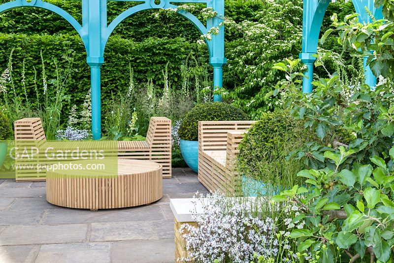 Wooden table and benches with blue metal arches and granite cobbles and clipped yew balls in planters - The Sir Simon Milton Foundation Garden: '500 years of Covent Garden' - RHS Chelsea Flower Show 2017 - Designer: Lee Bestall - Sponsor: Capco Covent Garden
