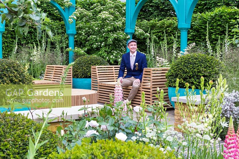 Designer Lee Bestall sitting on one of the wooden slatted benches on the garden with softly clipped yew balls  and roses featured in the surrounding planting. Cornus kousa behind - The Sir Simon Milton Foundation Garden: '500 years of Covent Garden' - RHS Chelsea Flower Show 2017 - Designer: Lee Bestall - Sponsor: Capco Covent Garden