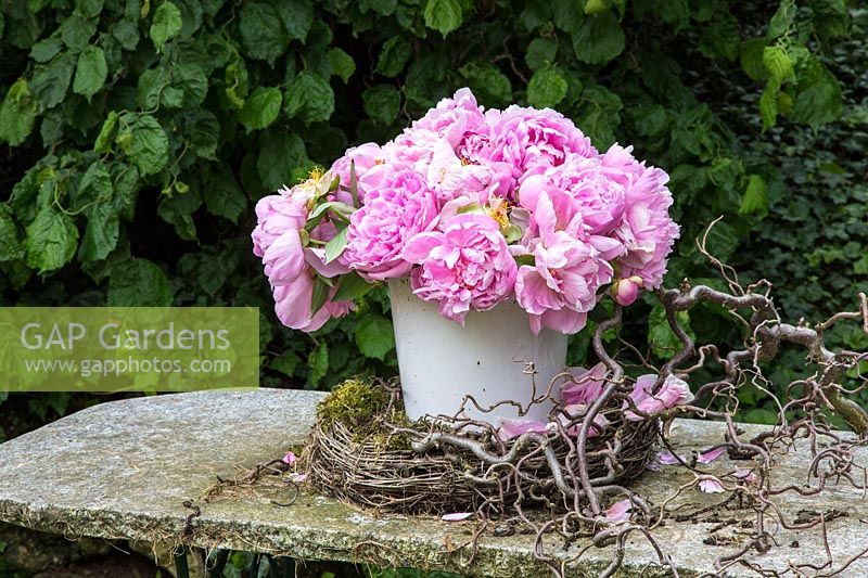 Bunch of pink Peonies in a white container on a stone table with a wreath and some hazel twigs