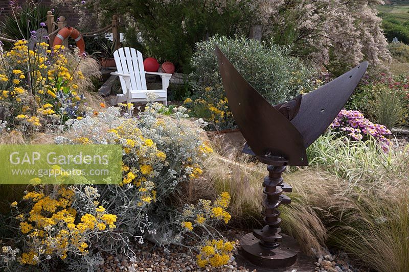 Seaside themed front garden with white Adirondack chair, planted with coastal plants, Cineraria 'Silver Dust', Stipa tenuissima, Erigeron 'Pink Jewel' and Tamarisk tetandra flowering behind. Decorated with driftwood sculptures,  lobster pots, fishing floats and an old rusted propeller