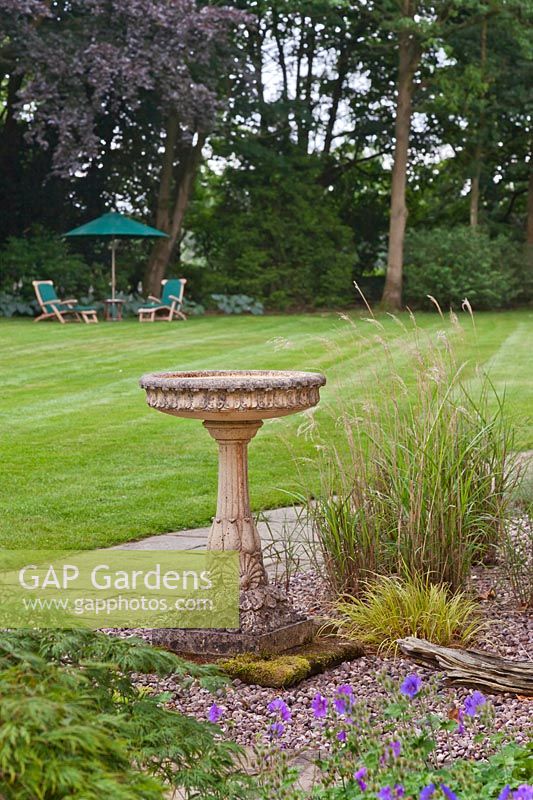 Traditional pedestal stone bird bath in front of lawn with steamer chairs and parasol - June, Cheshire