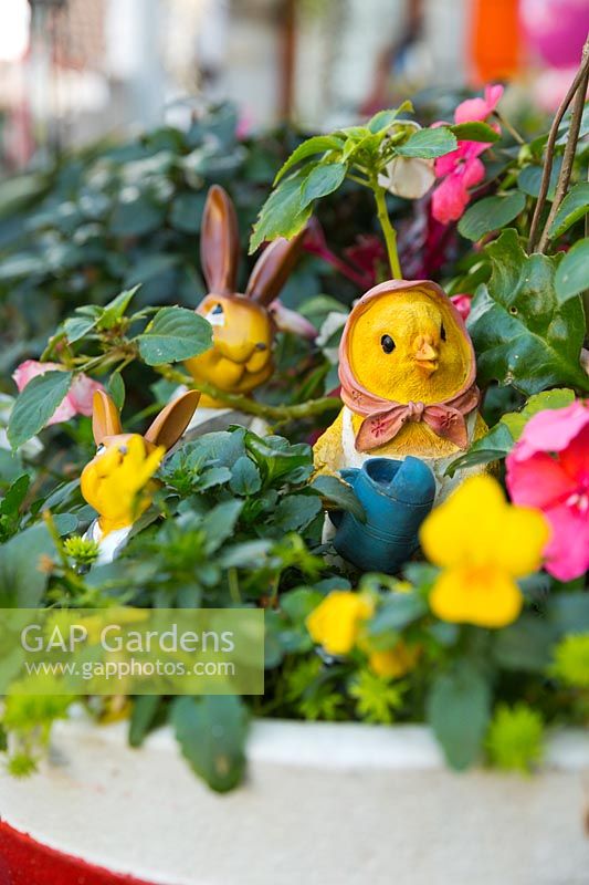 A pot with yellow Violas, pansies, pink Impatiens, two rabbits and a chick with yellow faces and decorations in the pot.