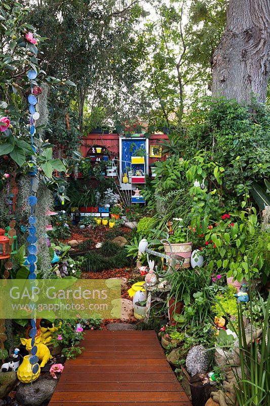 View from the lower part of the garden to the back fence, featuring an irregularly shaped timber deck, an eclectic collection of pots, garden ornaments, toys and shade loving plants.