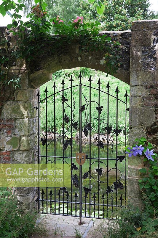 Wrought iron decorative gate with vine and grape motif set in ancient stone and flint wall, opening out to river. Clematis 'Blue Moon' on right.