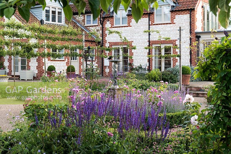 A modern extension, seen over a formal sunken parterre planted with Roses, Salvias, hard Geraniums, Astrantias and Catmint.