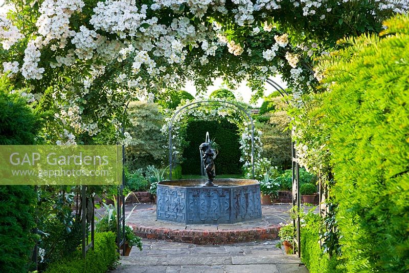 Small raised pond with 'Cherub' fountain, white delphiniums and metal arch with 'Rose' 'Wickwar'.
