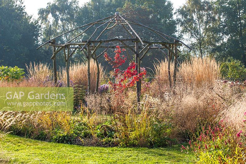 Rustic gazebo of coppiced ash and hazel surrounded by grasses such as Calamagrostis x acutiflora 'Karl Foerster'