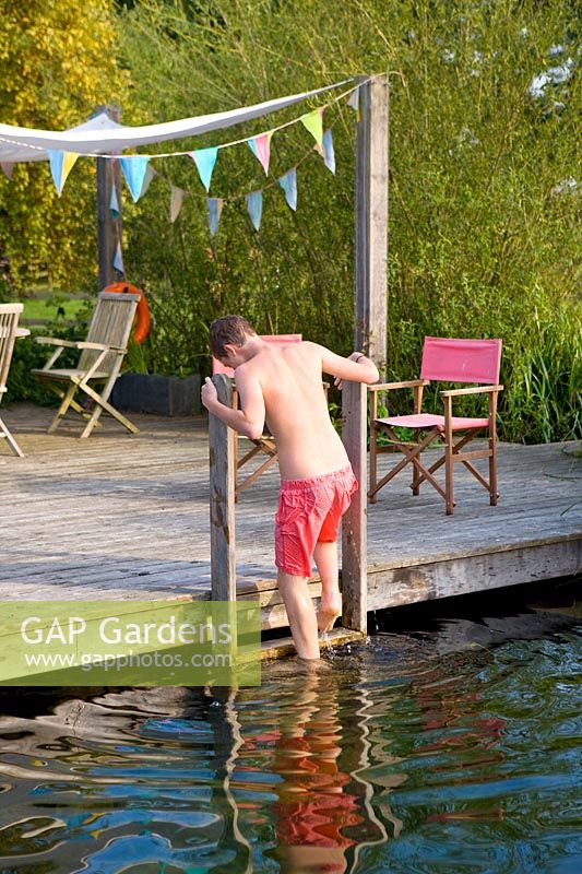 Boy gets out of 'natural' swimming pool by climbing up ladder attached to wooden deck seating area with shade sail

 