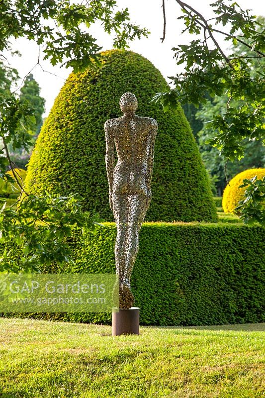 Sculpture of a figure in stainless steel on a lawn with topiary backdrop
