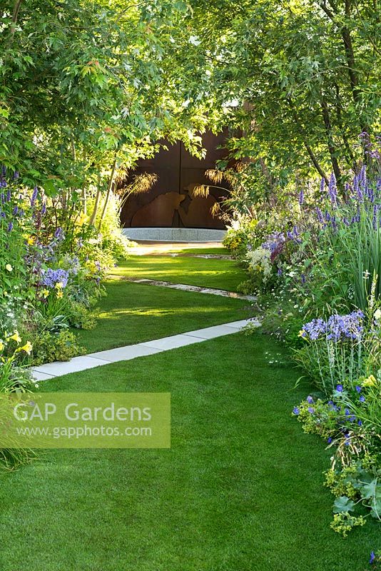White path crossing a lawn diagonally, with borders of trees and perennials. The A Dogs Life garden, RHS Hampton Court Show