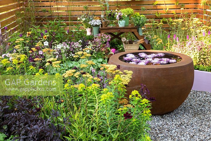 Small summer garden with flowering perennials and round water bowl with water lilies. Surrounded by wooden panel fencing