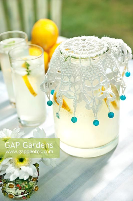 Jug of lemonade with crocheted beaded jug cover, drink tumblers, lemons and small vase of cut flowers