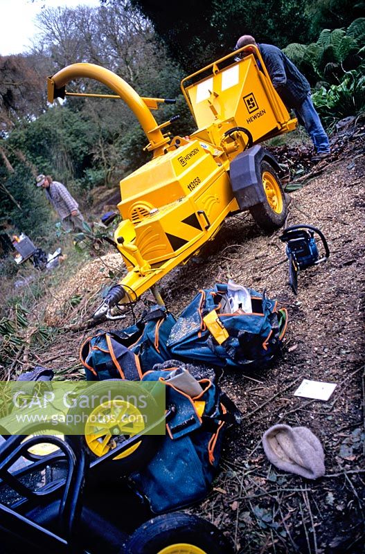 Large petrol engined garden chipper being used to clear overgrown garden Range of tools including wheelbarrow chainsaw