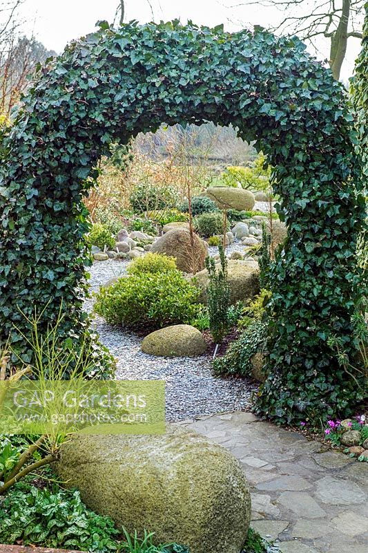 Arch of Hedera (ivy) over pathway leading to a garden of shrubs large boulders in John Massey's garden at Ashwood Nurseries