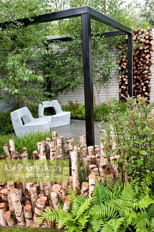 The Naturally Fashionable Garden designed by NDG+. Seating area with metal mesh seats, black pergola, birch logs & birch trees