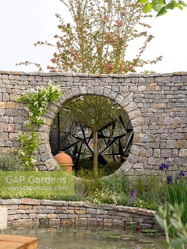 The Harmonious Garden of Life garden with a dry stone wall complete with large circular hole with view of the tree and background of reflective material bouncing light back into the garden.- Designer: Laurélie de la Salle - Sponsor: Mr Robert and Mrs Sue CawthornMargheriti Piante, Italy