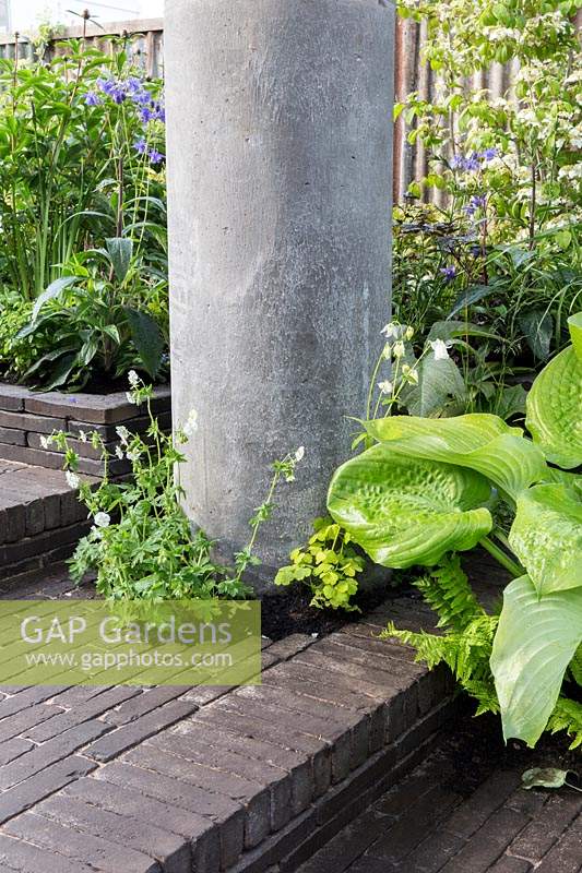 The Silent Pool Gin Garden, view of sunken garden with hostas against recycled concrete pillar, clay brick paving and corrugated metal fence – Designer: David Neale - Sponsor: Silent Pool Gin