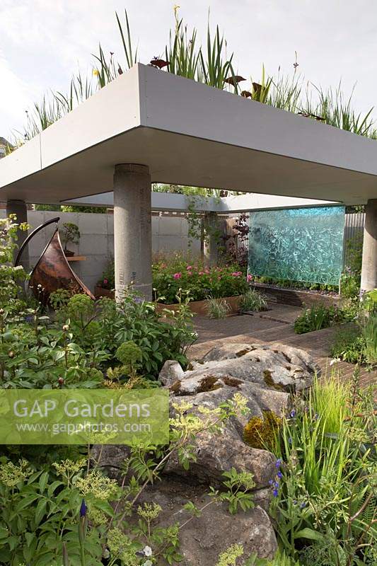 The Silent Pool Gin Garden, view of sunken garden with glass wall water feature, corrugated metal fencing, recycled concrete pillars supporting a roof with water garden, old limestone boulders,  clay brick wall and paving,  copper swing chair, roses, grasses – Designer: David Neale - Sponsor: Silent Pool Gin