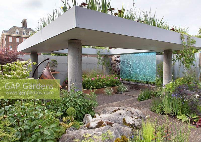 The Silent Pool Gin Garden, view of sunken garden with glass wall water feature, corrugated metal fencing, recycled concrete pillars supporting a roof with water garden, old limestone boulders,  clay brick wall and paving,  copper swing chair, roses, grasses – Designer: David Neale - Sponsor: Silent Pool Gin