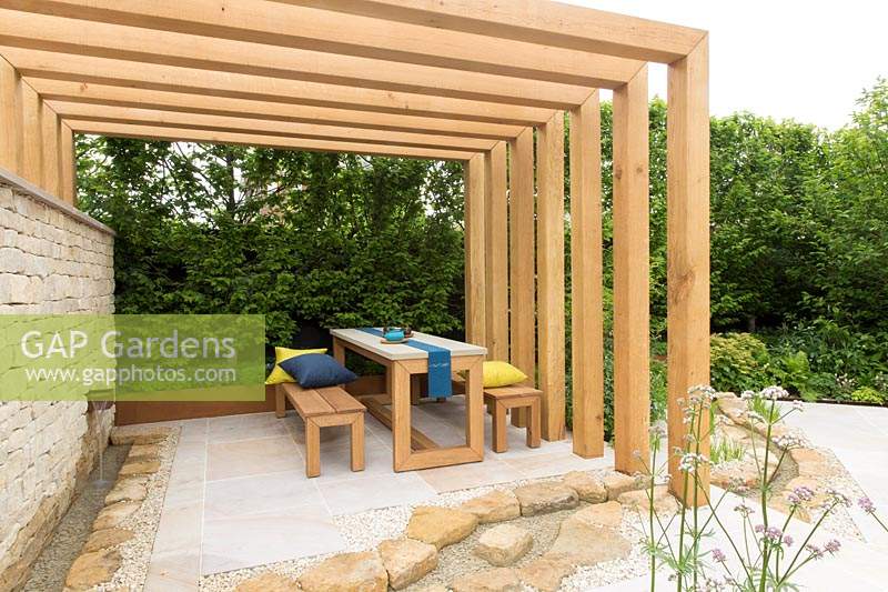 The Kampo No Niwa Garden, view of wood pergola with wooden table and bench seats and cushions, stone patio with water feature rill, dry stone wall with waterfall – Designer: Kazuto Kashiwakura and Miko Sato- Sponsor: Kampo No Niwa 300 sponsors. RHS Chelsea Flower Show 2019.