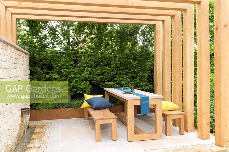 The Kampo No Niwa Garden, view of wood pergola with wooden table and bench seats and cushions, stone patio with water feature rill, dry stone wall with waterfall – Designer: Kazuto Kashiwakura and Miko Sato- Sponsor: Kampo No Niwa 300 sponsors. RHS Chelsea Flower Show 2019