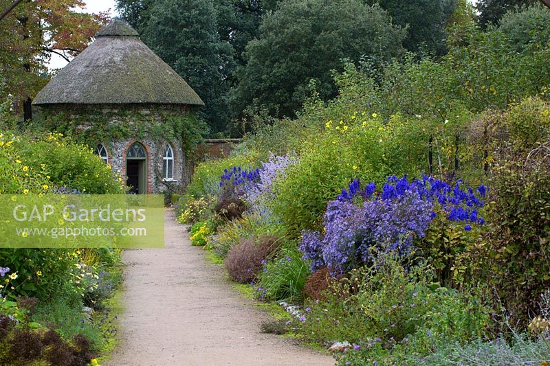 Double border of late-flowering perennials either side of a path leading to thatched building