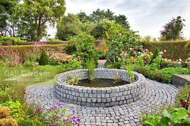 Circular raised pond made of stone setts with marginal planting and formal mixed borders.