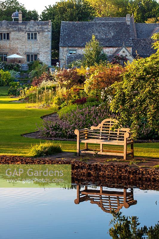 View across river coln to wooden lutyens bench, seat and borders beyond - Ablington Manor, Gloucestershire