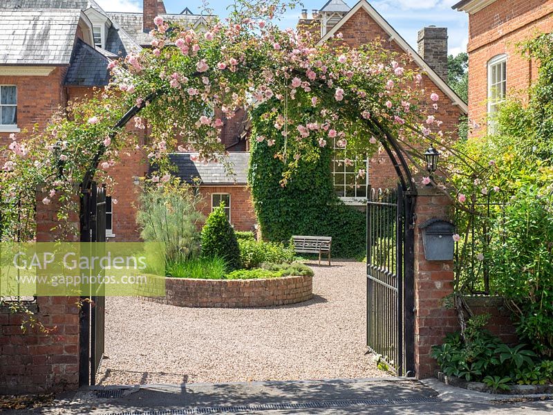 An iron arch covered with climbing pink Rosa, through which a raised herb bed can be seen.