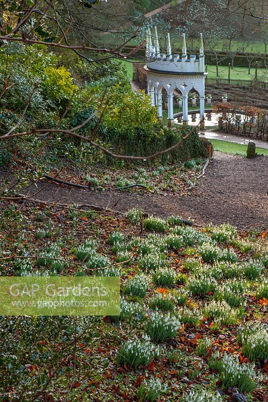 Path down slope beside snowdrops to a formal garden of exedra and pond