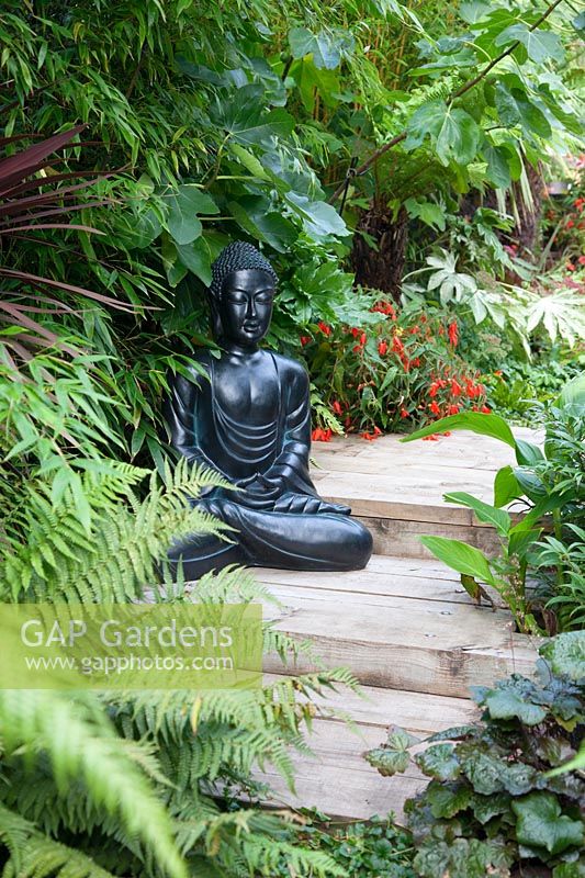 A black Buddha statue sits on a path built with railway sleepers amongst the lush foliage planting. 