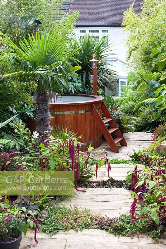 A wood fired hot tub sits on a wooden decked path surrounded by tropical planting. 
