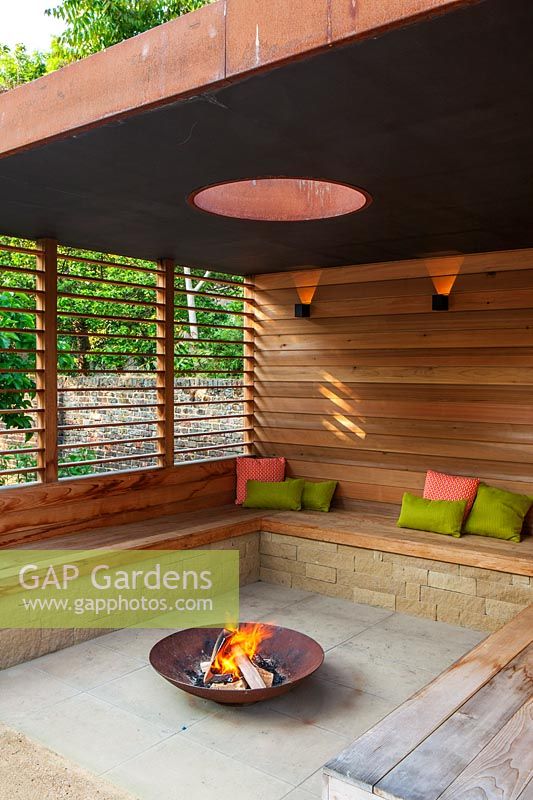 A contemporary pergola with fire pit and wooden bench seating. 
