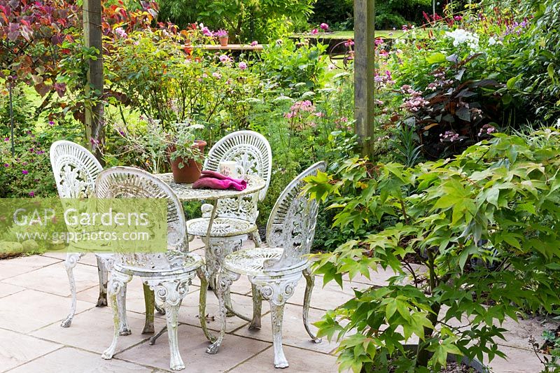 White metal garden table and chairs surrounded by shrubs and perennials. 

