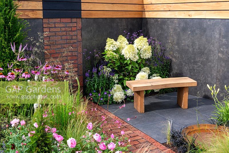 Brick path leading to a brick wall feature, wooden bench on slate tiles next to border with Hydrangea paniculata 'Phantom', Verbena bonariensis, Rosa 'Skylark' and Echinacea purpurea in foreground - A Place to Ponder, RHS Tatton Park Flower Show 2018